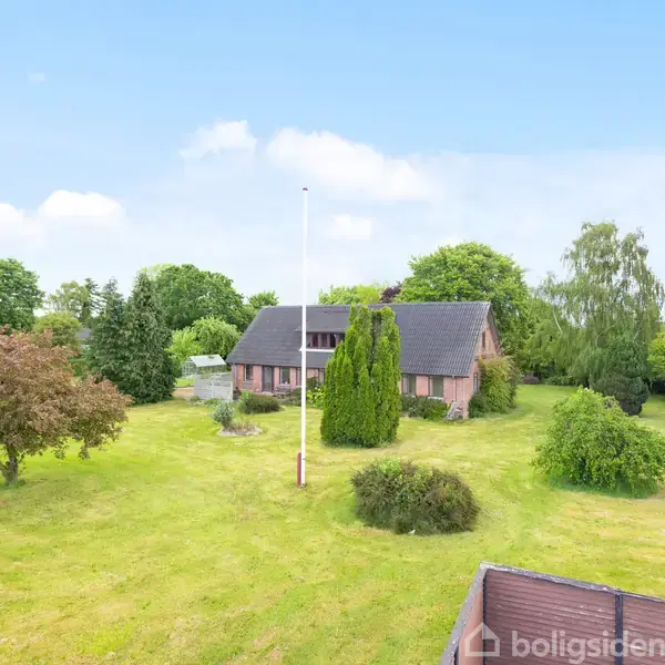Et rødt murstenshus med mørkt tag står i en stor, grøn have fyldt med træer og buske. En høj flagstang er placeret foran huset under en klar blå himmel.