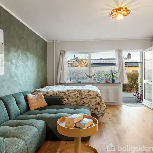 Grøn sofa med puder står mod en mintfarvet væg, ved siden af en seng med blomstret sengetæppe. Rummet har trægulv, vindue og glasdør ud mod en have. En rund bakke-bord med bøger står foran sofaen.