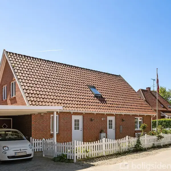Et rødt murstenshus med rødt tegltag og hvide vinduer står ved siden af en hvid stakit og carport. En hvid bil er parkeret i carporten. Grønne træer i baggrunden.