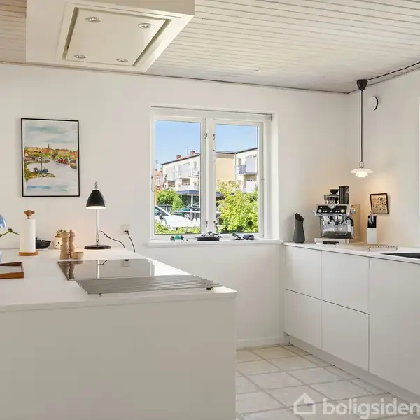 Køkken med hvide skabe, komfur på køkkenø, kaffemaskine og vindue med udsigt til bebyggelse. Lysindfald gennem stort vindue. Væghængt kunstværk til dekoration. Mørklagte lamper.