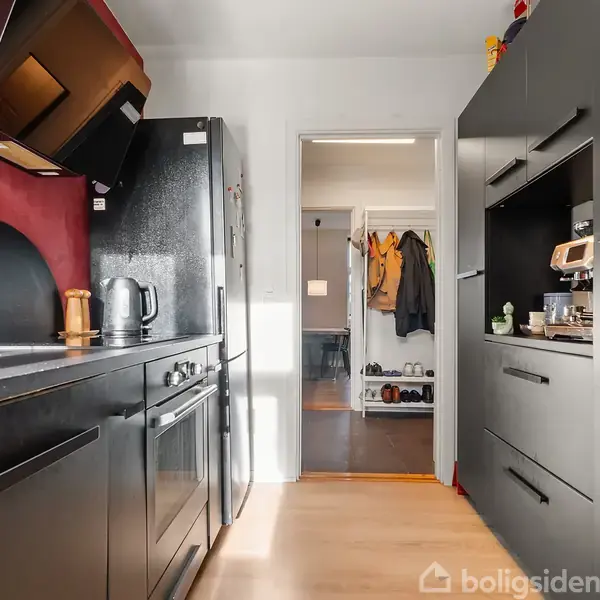 Køkken med sorte skabe på begge sider, køleskab og ovn til venstre. Glas- og trækopper står i venstre side under en rød væg. I gangen hænger jakker.