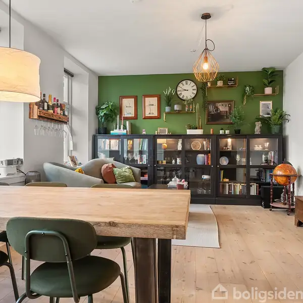 Et spisebord med grønne stole står i et lyst rum. En sofa og grøn væg med reol, klokke og planter skaber en hyggelig atmosfære. En globus står ved siden af.