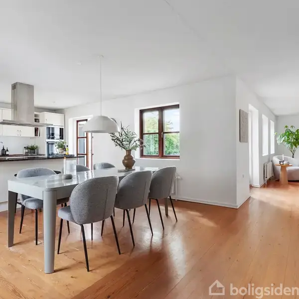 Spisestue med et gråt spisebord og seks stole, placeret ved siden af et åbent køkken. I baggrunden ses lys gennem vinduer, og gulvet er i lyst træ.