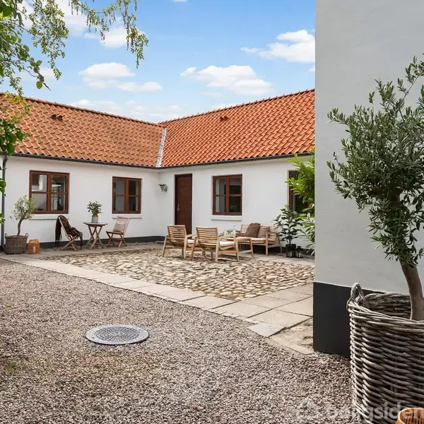 Et hvidt hus med rødt tegltag omkranser en gårdsplads med grusbelægning. Træmøbler og blomsterkrukker står i gårdspladsen. En blå himmel ses i baggrunden.