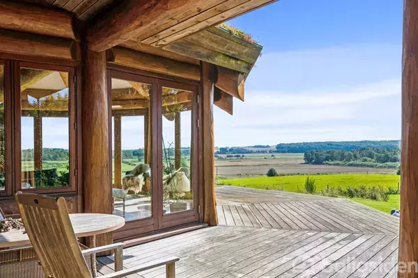 En træterrasse med et rundt bord og stol foran store glasvægge, der reflekterer landskab. Baggrunden viser en vid udsigt over marker og skove under en klar blå himmel.
