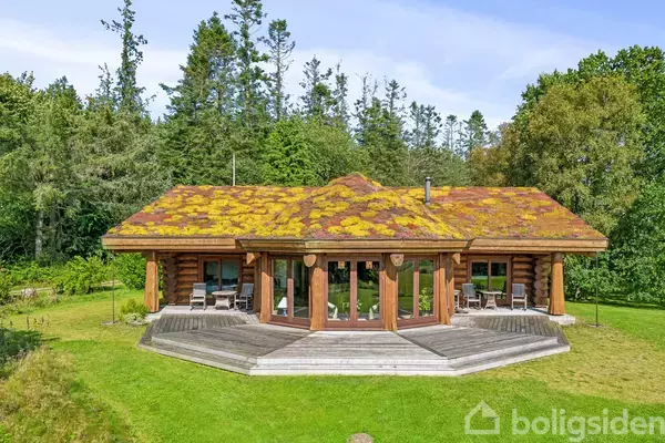 Et træhus med græstag står i en frodig have omgivet af høje træer. Huset har store vinduer og en terrasse med siddemøbler foran.