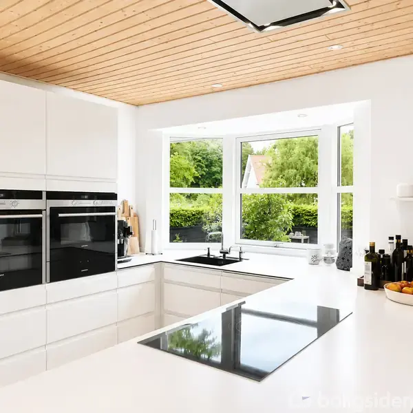Moderne køkken med hvide skabe og indbygget ovn. Kogeplader er monteret i en hvid bordplade. Store vinduer giver udsigt til grøn have. Flasker og frugtskål på bordet.