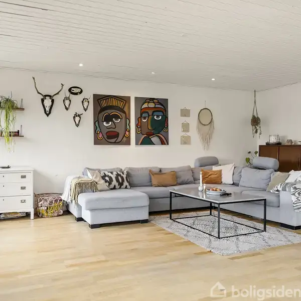 Grå sofa med puder står i stue. Vægdekorationer, inklusive malerier og hylder, hænger over sofaen. Et sofabord står på tæppe, omgivet af kommode og reol.