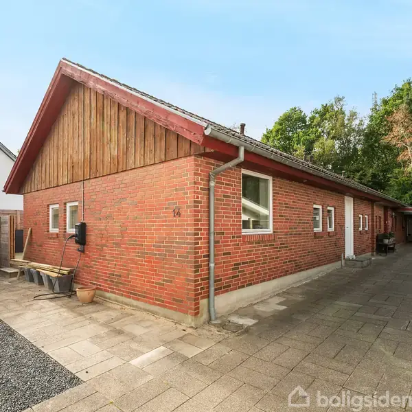 En rød murstensbygning ligger langs en flisebelagt indkørsel. Bygningen har et skråt tag med røde teglsten. Til venstre er en terrasse og i baggrunden ses træer.