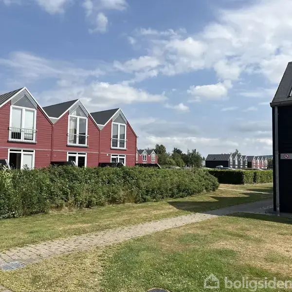 Rækkehuse med røde facader og sorte tage står på rad. De er omgivet af grønne hække og en brolagt sti under en blå, let skyet himmel.