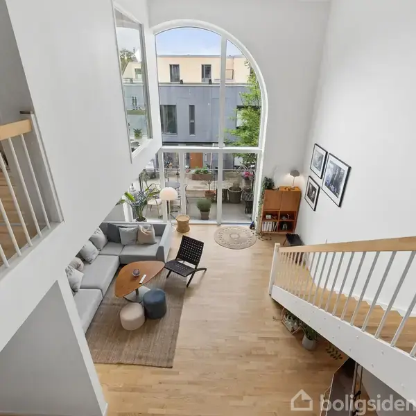 Stue set ovenfra viser sofa og stole ved sofabord, placeret på trægulv nær stor bueformet vindue. Etagen ovenfor har gelænder og reoler, og trappen fører nedad.