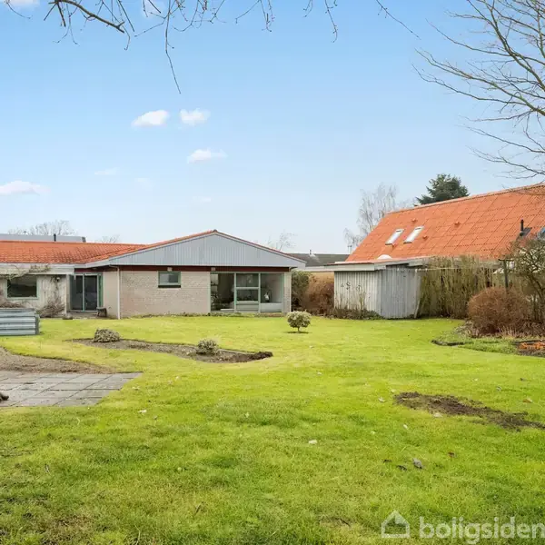 Et hus med rødt tag står i en have med grøn græsplæne og enkelte træer. Haven har en lille flisebelægning og et skur ved siden af huset.