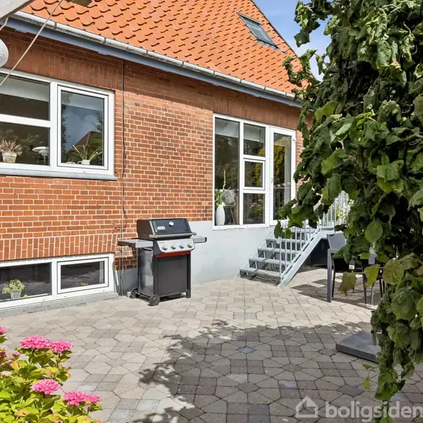 Grill står på en flisebelagt terrasse foran et rødt murstenshus. Trappe fører til en havedør. Til venstre ses lamper, og der er blomster i forgrunden.