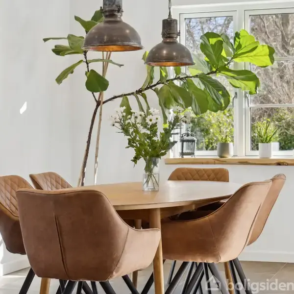 Et spisebord med brune stole står i et lyst rum. To lamper hænger over bordet, og en vase med grønne grene står i midten. Vindueskarm med planter ses i baggrunden.