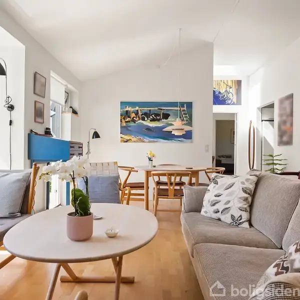 En stue med sofaer og et rundt bord med en hvid orkidé. I baggrunden et spisebord. Væggene er dekoreret med kunst, og store vinduer slipper lys ind.