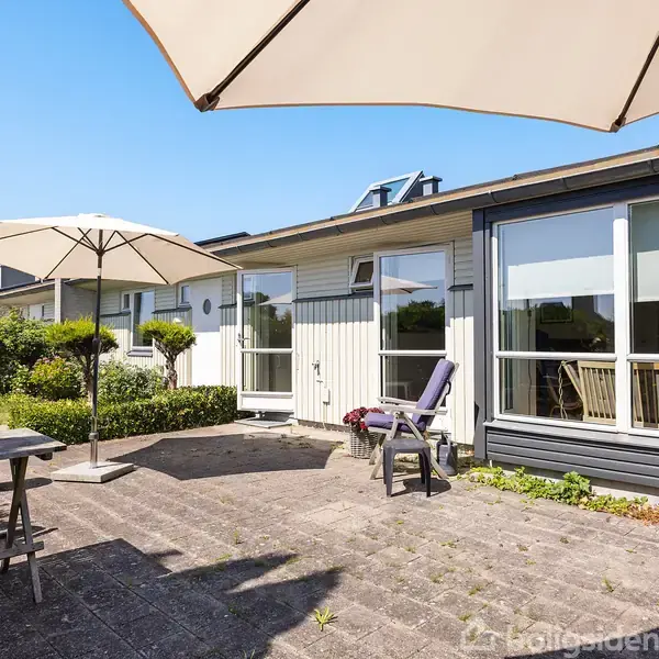 Terrasse med parasoller og havemøbler placeret på flisebelagt areal. I baggrunden ses en moderne bygning med store vinduer og hvid facade. Omkring er der buske og blå himmel.