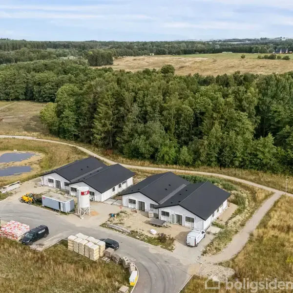 Tre hvide huse ligger ved siden af hinanden under opførelse. Omkring huse er en skov og åbne marker, mens byggeudstyr og materialer ligger tæt ved. En lille vej forbinder huse med det omkringliggende område.