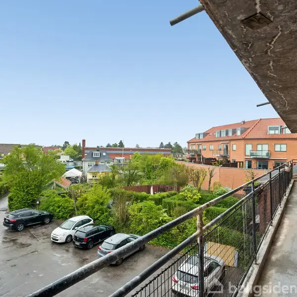 En betonaltan strækker sig langs en murstensbygning. Biler holder parkeret på en asfalteret plads omgivet af grønne træer og buske. I baggrunden ses flere tage og blå himmel.