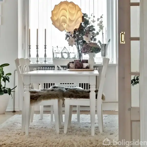 Et spisebord med hvide stole står under en dekorativ lampe. Bordet er dekoreret med blomster og lys, omgivet af et lyst rum med tæppe og planter ved vinduet.