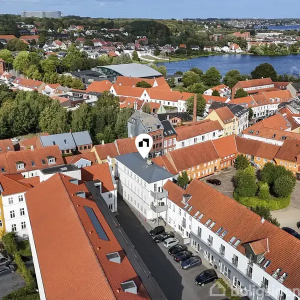 En by med røde tegltage set fra oven, hvor et hvidt hus er fremhævet med en markør. Biler er parkeret i gaderne. I baggrunden ses en sø og grønne skove.