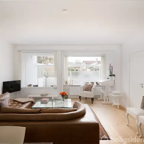 En stue med en stor brun lædersofa står i centrum. Sofaen er placeret omkring et glasbord. Store vinduer lader lys ind og afslører en lys og moderne indretning. Der er to hvide stole med pyntepuder og et lille arbejdsområde ved vinduet. Malerier hænger på væggen.