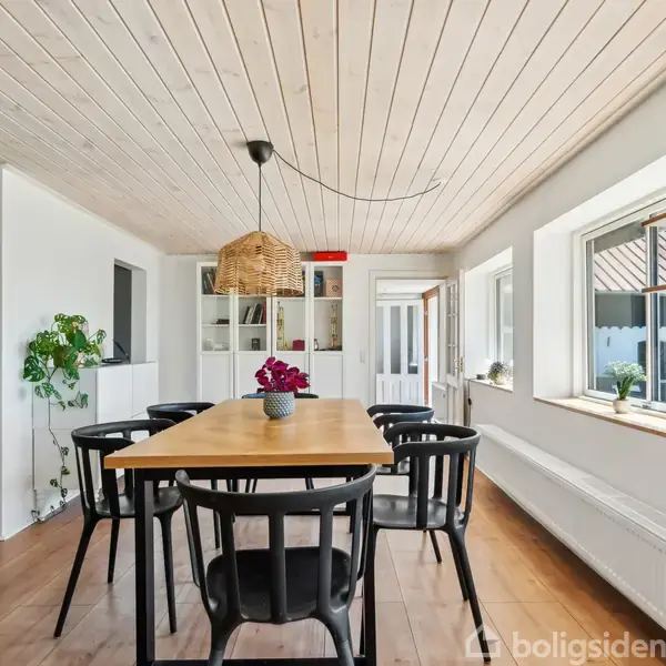 Et spisebord med sorte stole står i et lyst rum. Rummet har trægulv, lofter og store vinduer med dekorerede hylder. Plant og dekorationer skaber en hyggelig atmosfære.