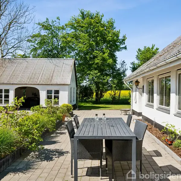 Et træbord med stole står på en flisebelagt terrasse. I baggrunden er der to hvide bygninger med store vinduer, omgivet af grønne træer og en blomstrende have.