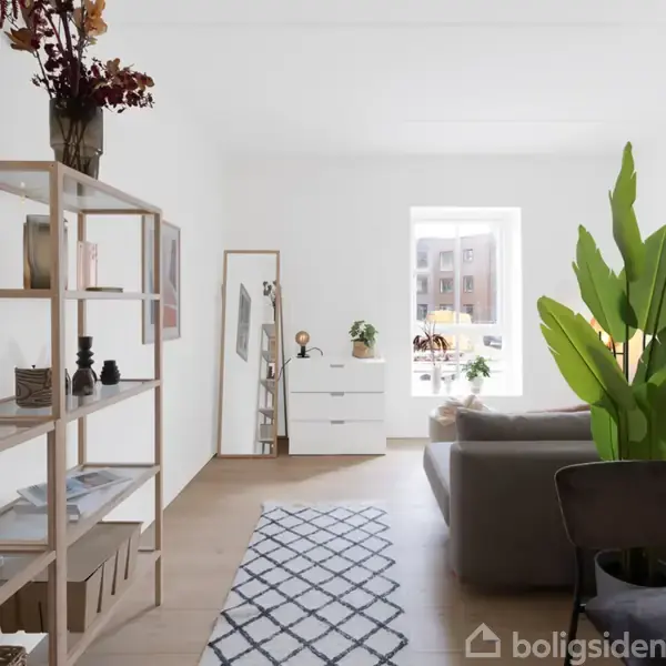 Reol med bøger og dekorationer står i en lys stue. Sofaen vender mod et vindue, planter skaber en grøn atmosfære, og et tæppe dækker lyst trægulv.