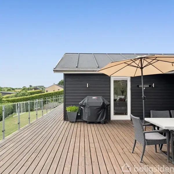 Stor træterrasse med et spisebordssæt under en parasol. En grill står ved den mørke husvæg. I baggrunden ses et grønt landskab og en blå himmel.