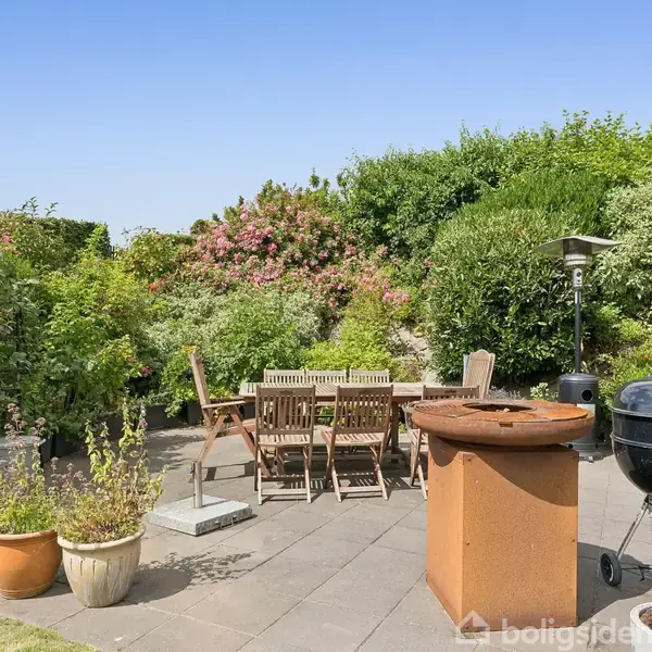 Terrasse med flisebelægning, hvor et rundt havebord og stole står omgivet af grønne buske og blomster. En grill er placeret til højre, og blomsterkrukker pryder området.