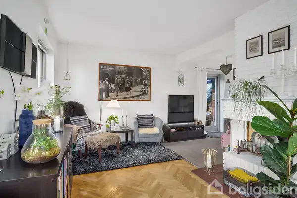 Stue med sofa og stol ved siden af et rundt bord, der bærer en lampe. Et stort billede hænger på væggen. Rummet er lyst med træ- og tæppegulv og moderne indretning.