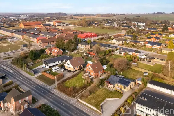 En række parcelhuse står stille langs en rolig vej i en forstad, omgivet af haver, træer og marker. Blandet beboelses- og landbrugsområde i baggrunden.