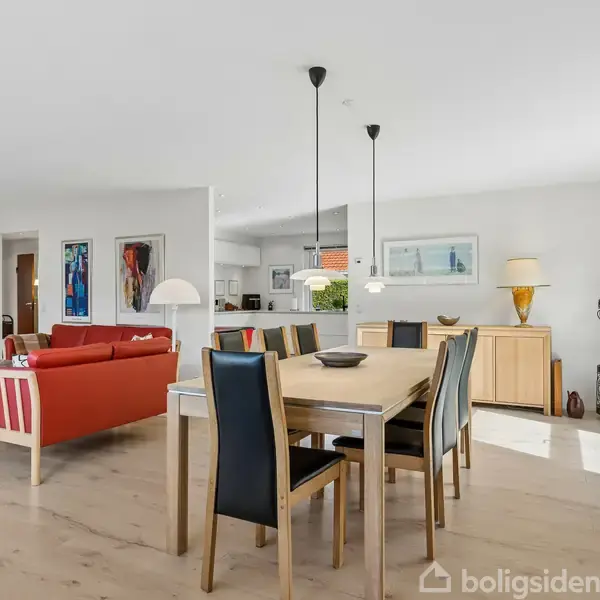 Spisebord med seks stole står midt i en lys stue. En rød sofa står bagved. Rummet er veloplyst af store vinduer, og der hænger lamper over bordet. Der ses kunst på væggene.
