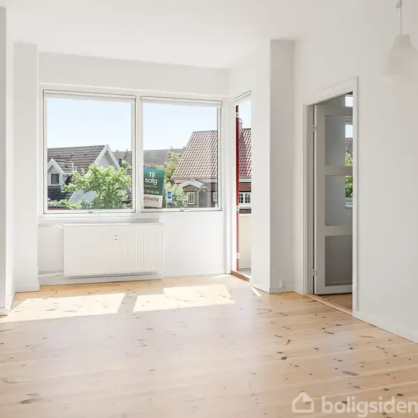 Et lyst rum med trægulv og hvide vægge, store vinduer med udsigt til nabohuse, en dør til en balkon og sollys der strømmer ind.