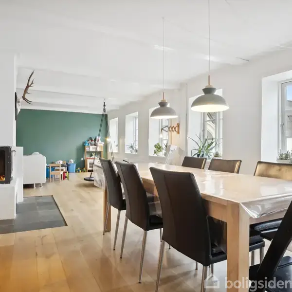 Spisebord med stole omkring i en moderne spisestue. Bordet står på et lyst trægulv nær store vinduer, mens pejsen til venstre er tændt, hvilket skaber en hyggelig atmosfære.