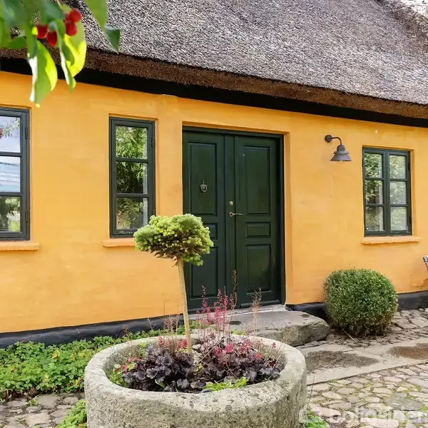 En gulmalet bindingsværkshus ses med grønne vinduer og dør. Foran huset er en stenplantekumme med planter. En træbænk står ved siden af, omgivet af en brostensbelægning og grønne omgivelser.