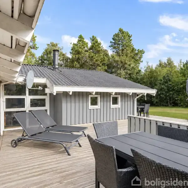 To solsenge står stille på en træterrasse foran et gråt sommerhus med hvide vinduesrammer. I baggrunden ses græsplæne og træer under en blå himmel med skyer.