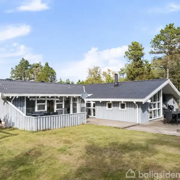 Gråmalet træhus med sort tag står på en grøn græsplæne. En terrasse med spisebord og stole ligger på højre side. Træer omkranser området under en blå himmel.