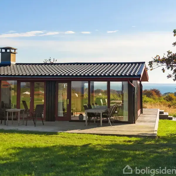 Sommerhus med glasvægge står stille i en frodig have. Græsplæne fører til terrasse, hvor havet skimtes i baggrunden under en klar, blå himmel.