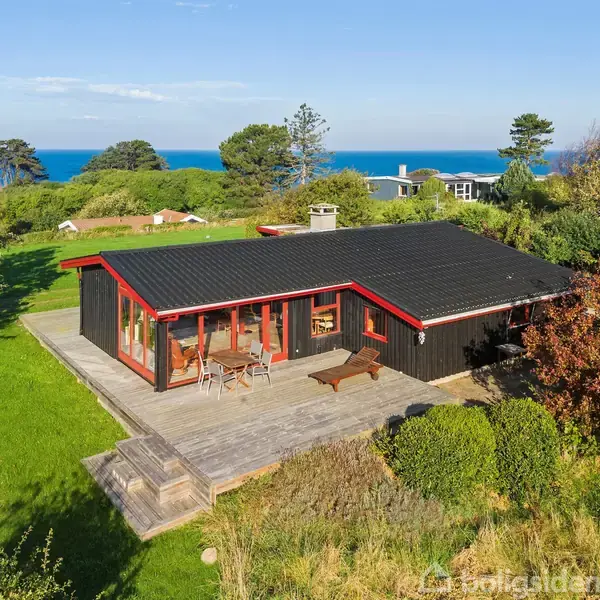 Sommerhus med sort træfacade og røde detaljer står på en græsplæne med terrasse, omgivet af buske og træer. I baggrunden ses havet og flere mindre huse.
