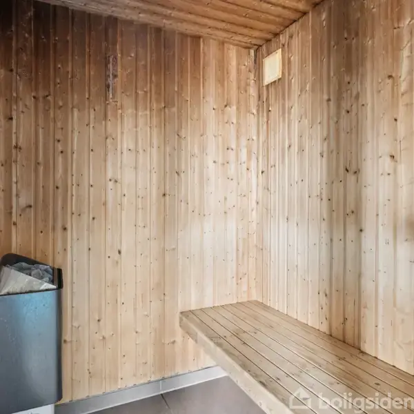Sauna med træbeklædte vægge rummer en bænk til at sidde på. Et elektrisk varmeapparat med sten står i hjørnet. Æstetisk enkelhed i et afslappende rum.