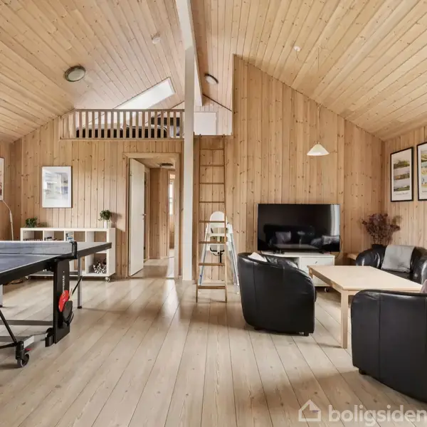 Stue med træinteriør har bordtennisbord og lænestole. TV står på et møbel ved siden af en sofa. Væggene har kunsttryk, og rummet er lyst op af store vinduer.