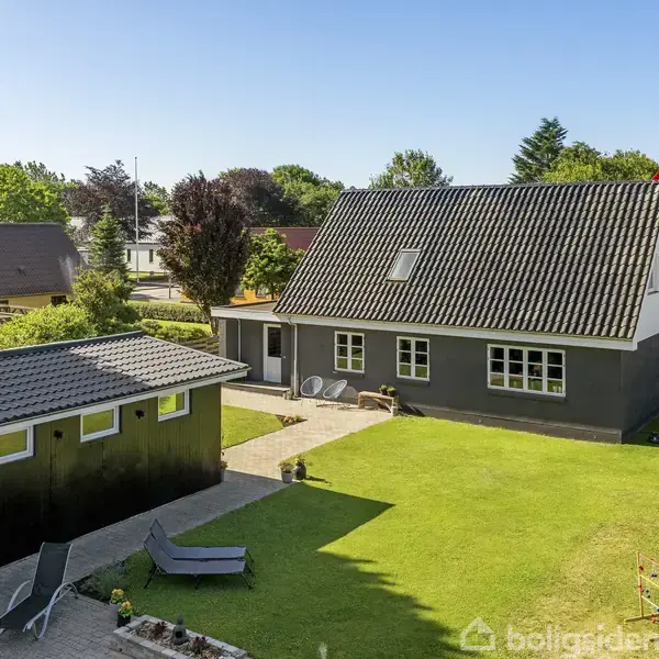 Et sort-hvidt hus med mansardtag står på en velplejet græsplæne. Husets have har fliser og liggestole. Omkring er der flere huse og grønne træer.
