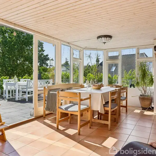 Et lyst rum med et spisebord og stole ved et stort vinduesparti. Bordet er dækket med en dug, og planter står i hjørnerne. Udendørs ses en terrasse med havemøbler.