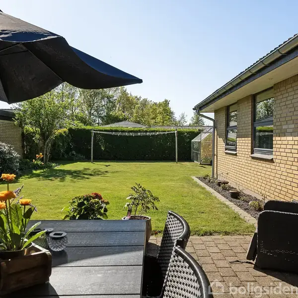 En terrasse med et bord og stole er placeret ved siden af et gult murstenshus. Bordet har en sort parasol og en blomsterkrukke med orange blomster. Græsplæne og hæk i baggrunden.