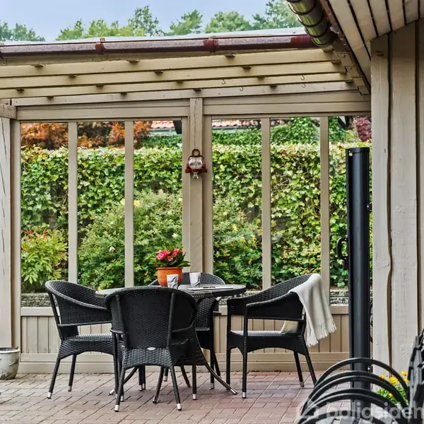 Et bord med fire sorte stole står under en overdækket terrasse. En plante i potte og en dug pryder bordet. Baggrunden viser en hæk og en plæne.