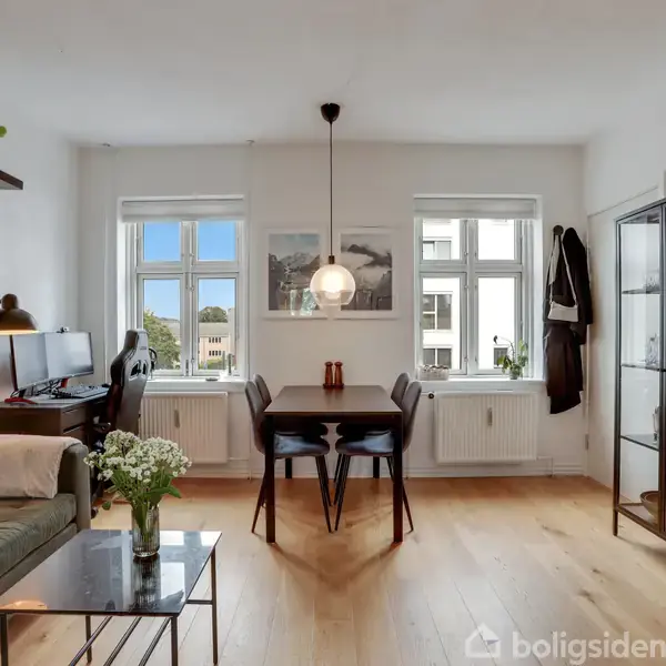 Et spisebord med fire stole står foran to vinduer. Til venstre er en grøn sofa og et skrivebord med lampe. Til højre er et vitrineskab. Rummet har trægulv.
