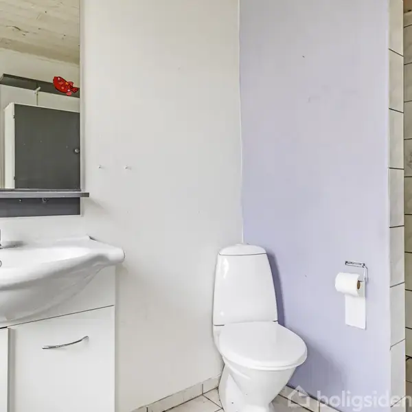 Et badeværelse med hvidt toilet og håndvask. Håndvasken har et indbygget skab. Toilettet er ved siden af en lyselilla væg. Der ses et spejl over håndvasken.