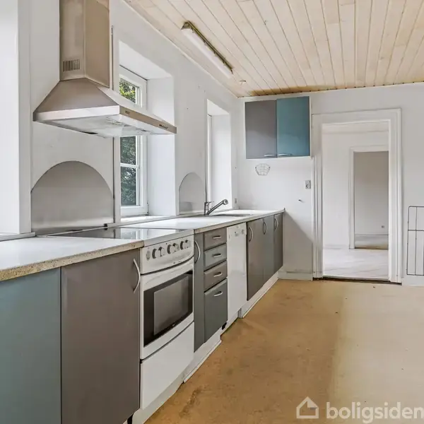 Køkken med grå og blå skabe, komfur og emhætte på en lang bordplade. Stort vindue til venstre giver udsigt til grønt område. Værelset har trægulv og lyst loft.