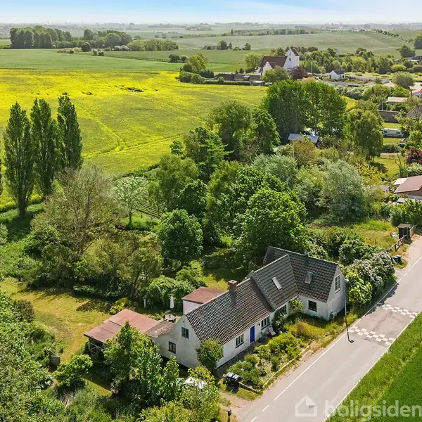 Hus omgivet af frodig have står stille langs vej i naturskøn landlig landskab med marker og træer. En landsby ses i baggrunden under en klar himmel.
